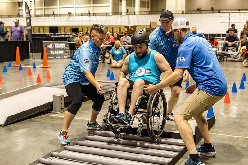 A man in a wheelchair wearing a helmet is assisted by three individuals as he navigates a slalom course indoors. The setting resembles a sports event, with cones and ramps visible. Spectators are in the background.