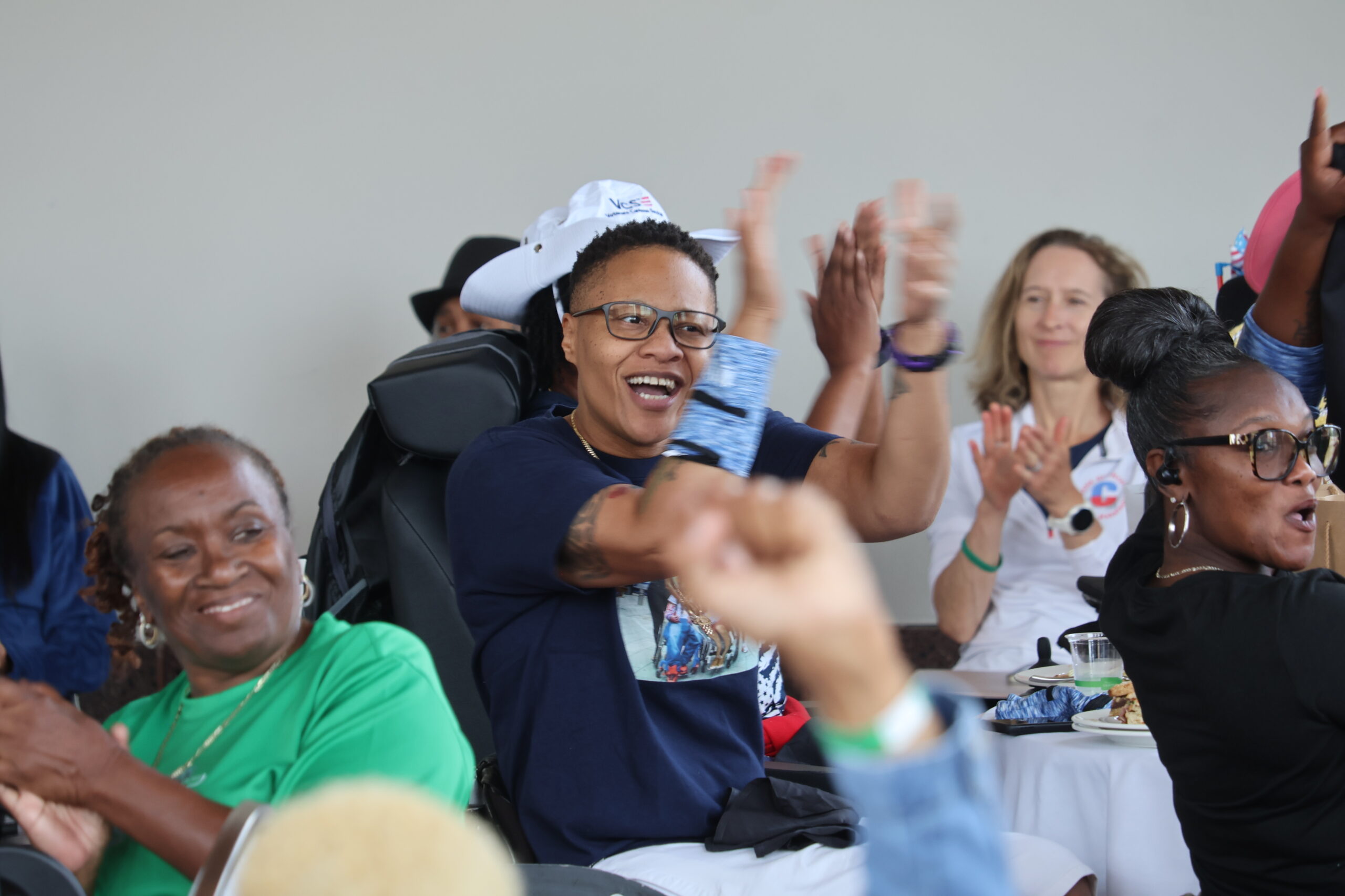 A group of people sitting and smiling, clapping their hands in celebration. One person in the center is especially enthusiastic, wearing glasses, a hat, and a navy blue shirt. The atmosphere is joyful and lively.