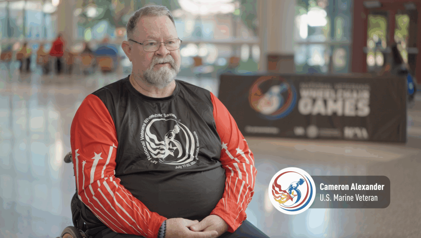 A man in a wheelchair wearing a red and black athletic shirt sits indoors. A sign in the background reads "National Veteran Wheelchair Games." Text on screen identifies him as Cameron Alexander, U.S. Marine Veteran.