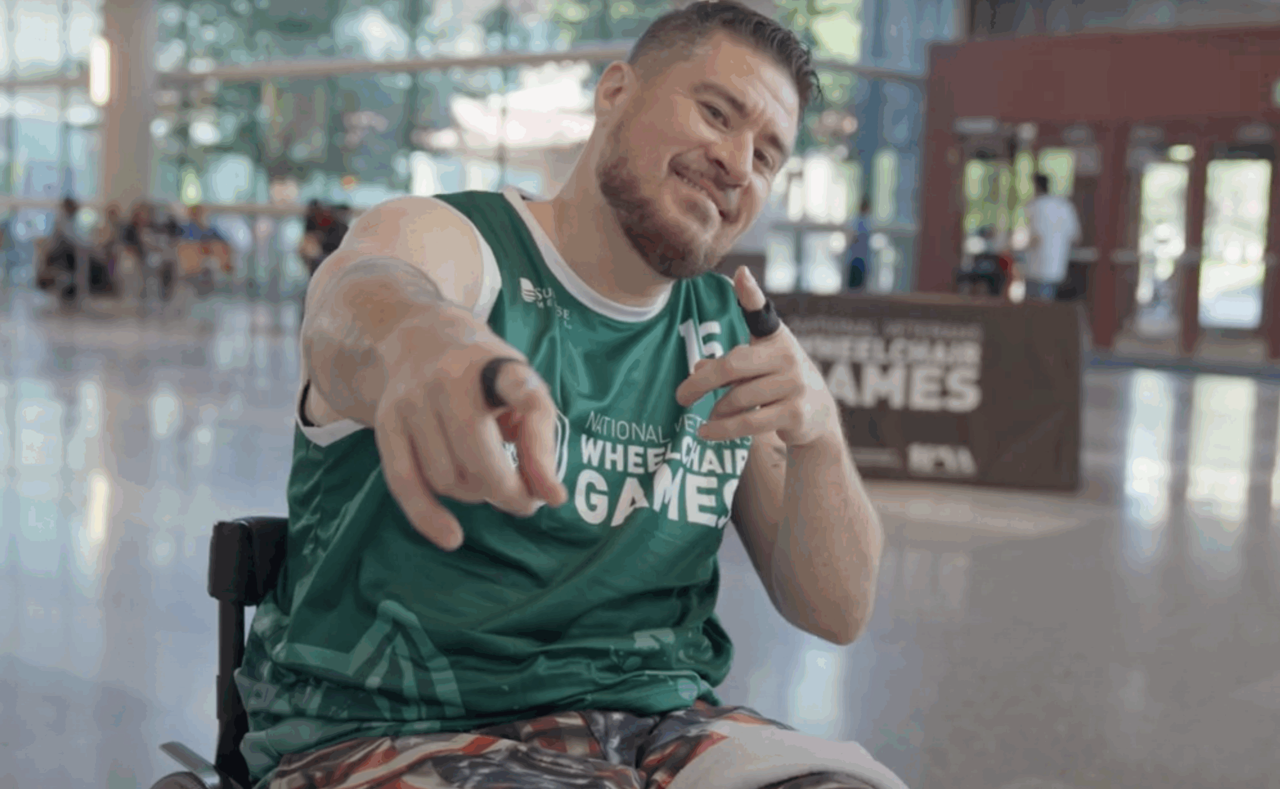 A smiling man in a green "National Veterans Wheelchair Games" jersey sits in a wheelchair indoors, pointing playfully at the camera. People and a games sign are visible in the background.