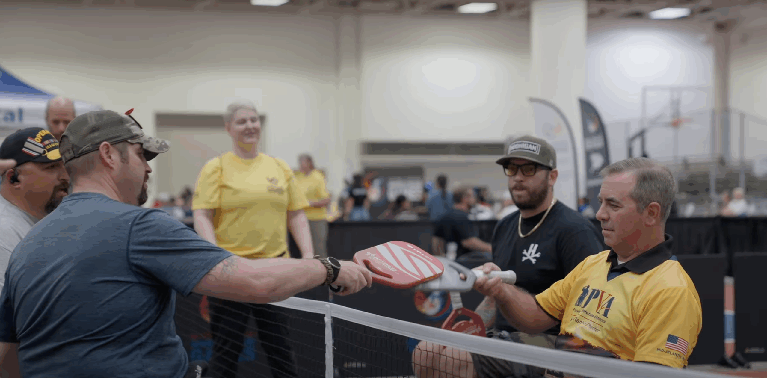Four men, two in yellow shirts and two in casual clothes, sit and play pickleball at an indoor event. One man in a yellow shirt uses a wheelchair. The net and paddles are visible; a woman in yellow stands in the background, smiling.