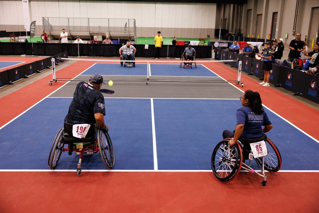 Two athletes in wheelchairs play a game of pickleball on an indoor court while spectators and other competitors watch from the sidelines.
