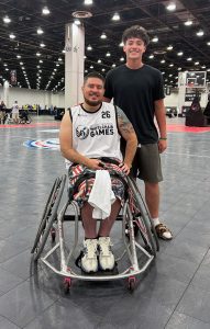 A man in a wheelchair wearing a "Wheelchair Games" jersey poses with a smiling teen in casual clothes on an indoor basketball court.
