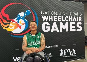 A smiling athlete in a wheelchair poses in front of a "National Veterans Wheelchair Games" banner. The athlete wears a green jersey and has visible tattoos on their arms.