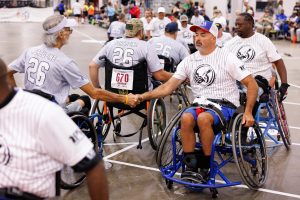 Two men in wheelchairs, wearing sports uniforms, shake hands on an indoor court. Other players in wheelchairs and spectators are visible in the background. The atmosphere is friendly and competitive.