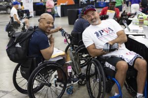 Two men in wheelchairs smile at the camera at an indoor event, one wearing a t-shirt and hat with "PVA" on it. There are other people and tables in the background.